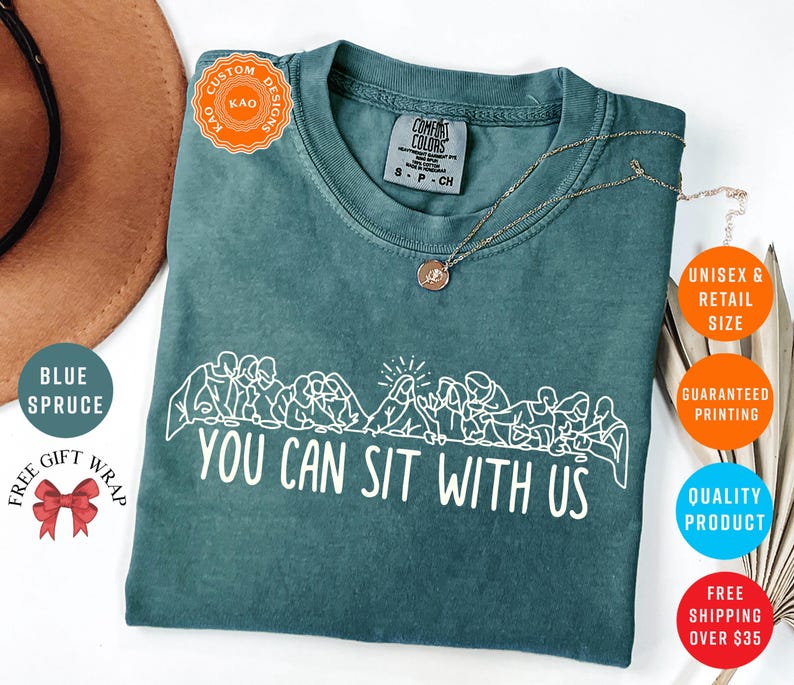 May include: Blue spruce Comfort Colors t-shirt with the text "YOU CAN SIT WITH US" in white. The shirt features a white graphic of people sitting. A gold necklace is draped on the shirt. The shirt is a unisex retail size.