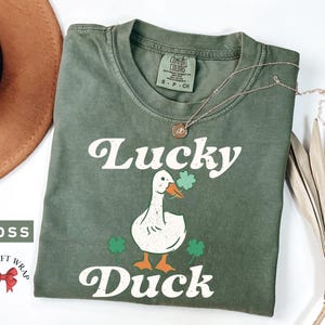Pu&ograve; includere: Una maglietta verde con un'anatra bianca che indossa un trifoglio e il testo "Lucky Duck" in un font retr&ograve;.