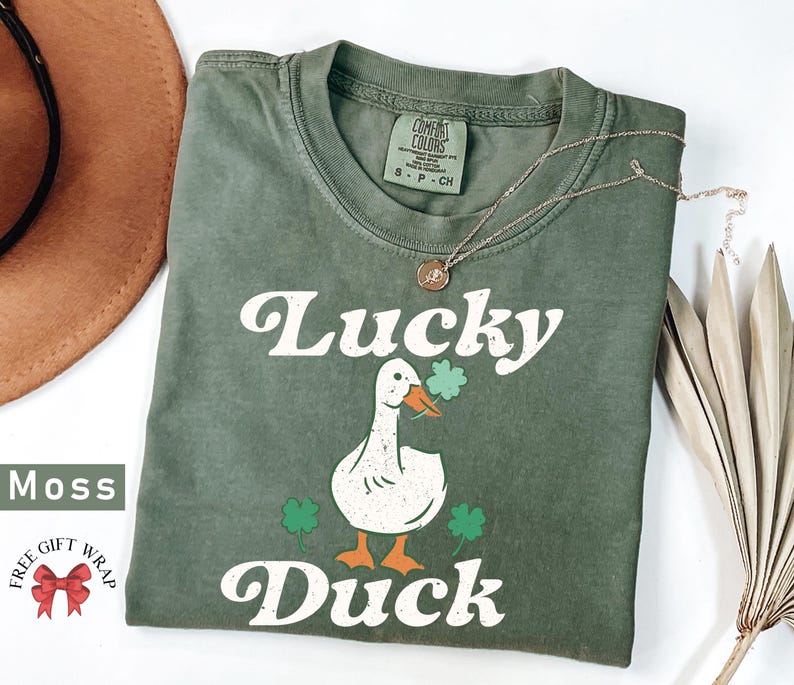 Pu&ograve; includere: Una maglietta verde con un'anatra bianca che indossa un trifoglio e il testo "Lucky Duck" in un font retr&ograve;.