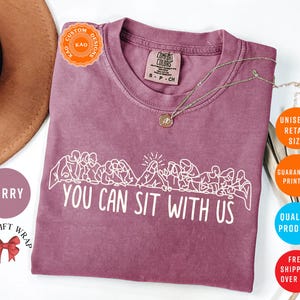 May include: Berry-colored t-shirt with the text "YOU CAN SIT WITH US" in white. The shirt features a crew neck and short sleeves. A gold necklace with a pendant is draped on the shirt. The shirt is a unisex retail size.