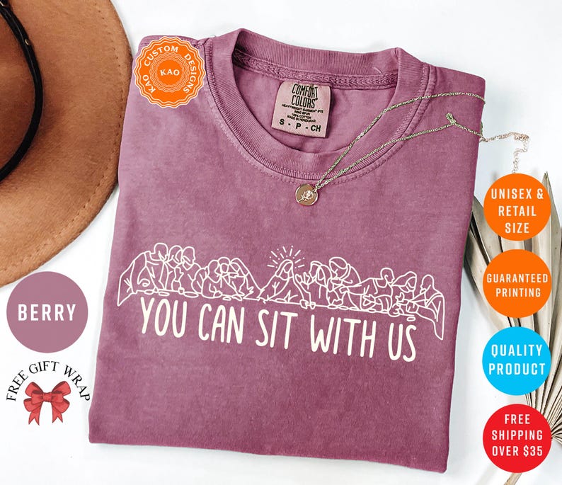 May include: Berry-colored t-shirt with the text "YOU CAN SIT WITH US" in white. The shirt features a crew neck and short sleeves. A gold necklace with a pendant is draped on the shirt. The shirt is a unisex retail size.