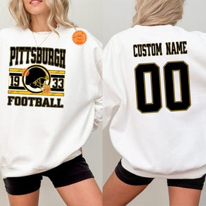 May include: White sweatshirt with a Pittsburgh Steelers football design. The front features the team name, helmet graphic, and the years 1933. The back has the option for a custom name and the number 00 in black and gold.