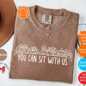 May include: Espresso-colored t-shirt with the text "YOU CAN SIT WITH US" in white. The shirt features a graphic of people sitting together. A silver necklace with a pendant is on the shirt.