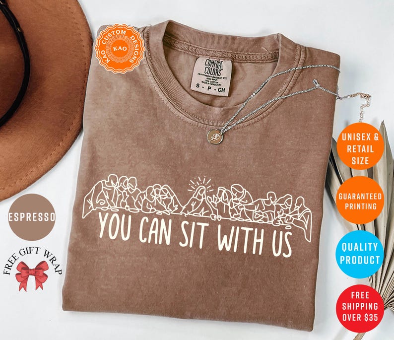 May include: Espresso-colored t-shirt with the text "YOU CAN SIT WITH US" in white. The shirt features a graphic of people sitting together. A silver necklace with a pendant is on the shirt.