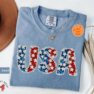 May include: A blue jean t-shirt with the word "USA" in red, white, and blue sequins. The shirt has a distressed look and is made by Comfort Colors. The text "Custom Designs Kao" is on a small orange tag.