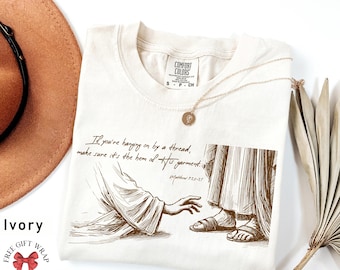 May include: Ivory t-shirt with a brown illustration of Jesus reaching out to a person. The text "If you're hanging on by a thread, make sure it's the hem of his garment." and "Matthew 9:20-21" are printed on the shirt.