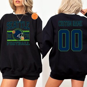 May include: Black sweatshirt featuring a Seattle football design with a helmet graphic, the year 1976, and the word "FOOTBALL". The back of the sweatshirt has the text "CUSTOM NAME" above the number "00".