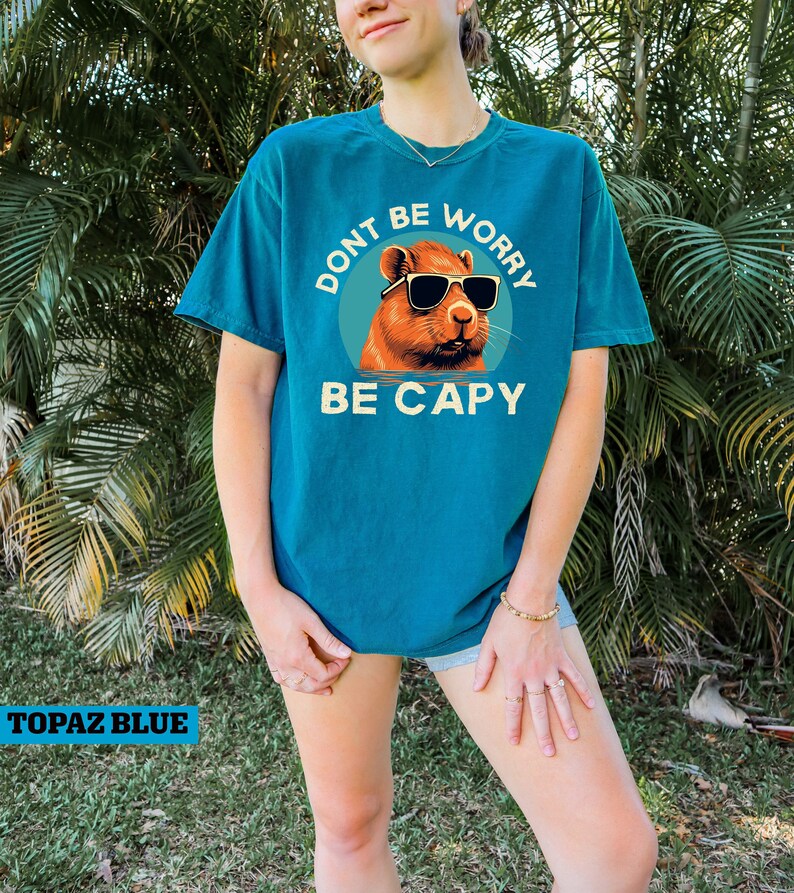 May include: A turquoise blue t-shirt with a graphic of a capybara wearing sunglasses. The graphic has the text "Don't be worry, be capy" around it.