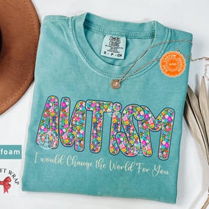 May include: Seafoam green t-shirt with the word "AUTISM" in a floral design. The text "I would Change the World For You" is below the word "AUTISM". A gold necklace and a brown hat are also in the image.