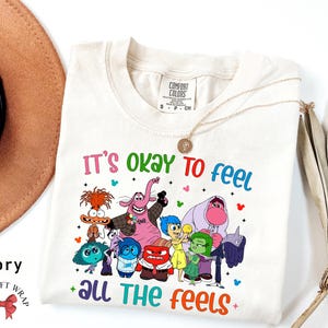 May include: Ivory t-shirt with a graphic of the characters from the movie Inside Out. The text "It's okay to feel all the feels" is printed on the shirt.