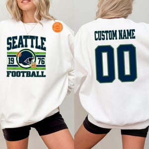 May include: White sweatshirt with "SEATTLE FOOTBALL" text and a football helmet graphic. The front design includes "1976". The back features "CUSTOM NAME" above the number "00" in navy blue with a green outline. Gildan brand.