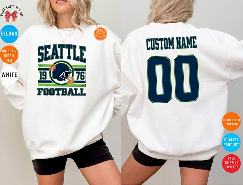 May include: White sweatshirt with "SEATTLE FOOTBALL" text and a football helmet graphic. The front design includes "1976". The back features "CUSTOM NAME" above the number "00" in navy blue with a green outline. Gildan brand.