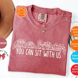 May include: Crimson-colored t-shirt with the text "YOU CAN SIT WITH US" in white. The shirt features a line drawing of people sitting together. The shirt is unisex and available in retail sizes.