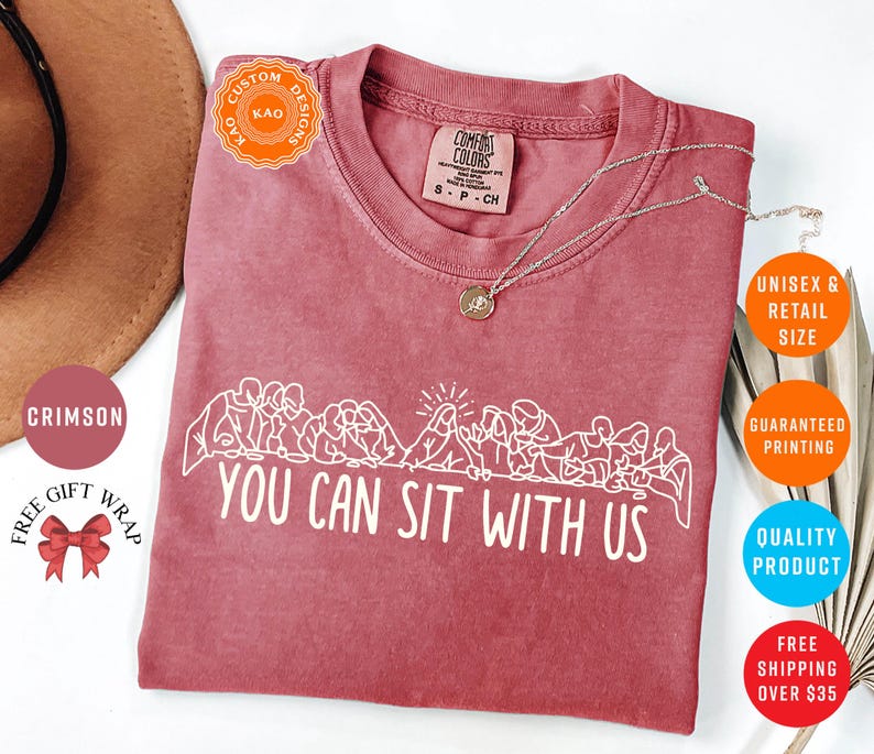 May include: Crimson-colored t-shirt with the text "YOU CAN SIT WITH US" in white. The shirt features a line drawing of people sitting together. The shirt is unisex and available in retail sizes.