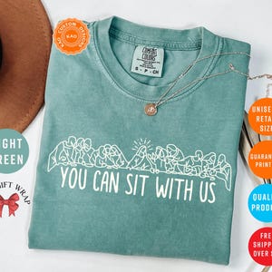 May include: Light green Comfort Colors t-shirt with the text "YOU CAN SIT WITH US" in white. The shirt features a graphic of people sitting together. A silver necklace is draped across the shirt.