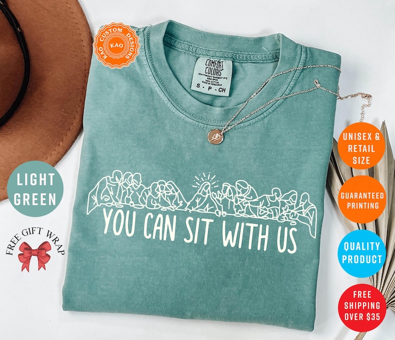 May include: Light green Comfort Colors t-shirt with the text "YOU CAN SIT WITH US" in white. The shirt features a graphic of people sitting together. A silver necklace is draped across the shirt.