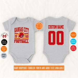 May include: A light gray baby bodysuit with a red and gold "Kansas City Football" design on the front, and a customizable name and number on the back. The bodysuit is available in multiple sizes.