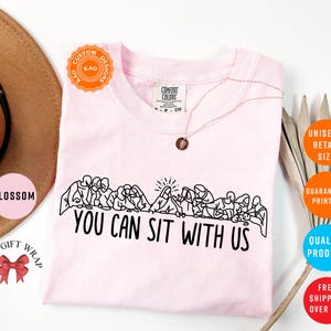 May include: A light pink t-shirt with a black graphic of people seated at a table, with the text "YOU CAN SIT WITH US". The shirt has a crew neck and short sleeves. A gold necklace is visible.