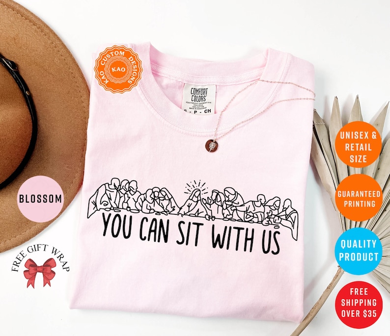 May include: A light pink t-shirt with a black graphic of people seated at a table, with the text "YOU CAN SIT WITH US". The shirt has a crew neck and short sleeves. A gold necklace is visible.
