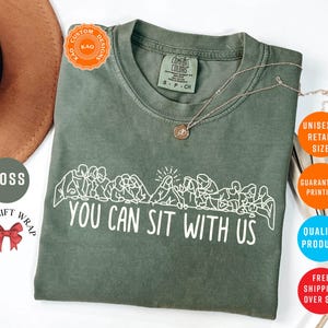 May include: A moss green t-shirt with the text "YOU CAN SIT WITH US" in white. The shirt features a graphic of people sitting together. A gold necklace is draped on the shirt. The shirt is a unisex retail size.