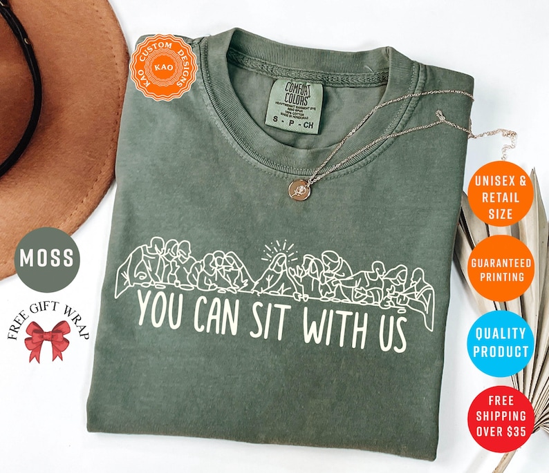 May include: A moss green t-shirt with the text "YOU CAN SIT WITH US" in white. The shirt features a graphic of people sitting together. A gold necklace is draped on the shirt. The shirt is a unisex retail size.