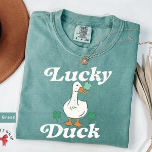 Pu&ograve; includere: Maglietta verde chiaro con un'anatra bianca che tiene un trifoglio e il testo "Lucky Duck" in un font retr&ograve;.