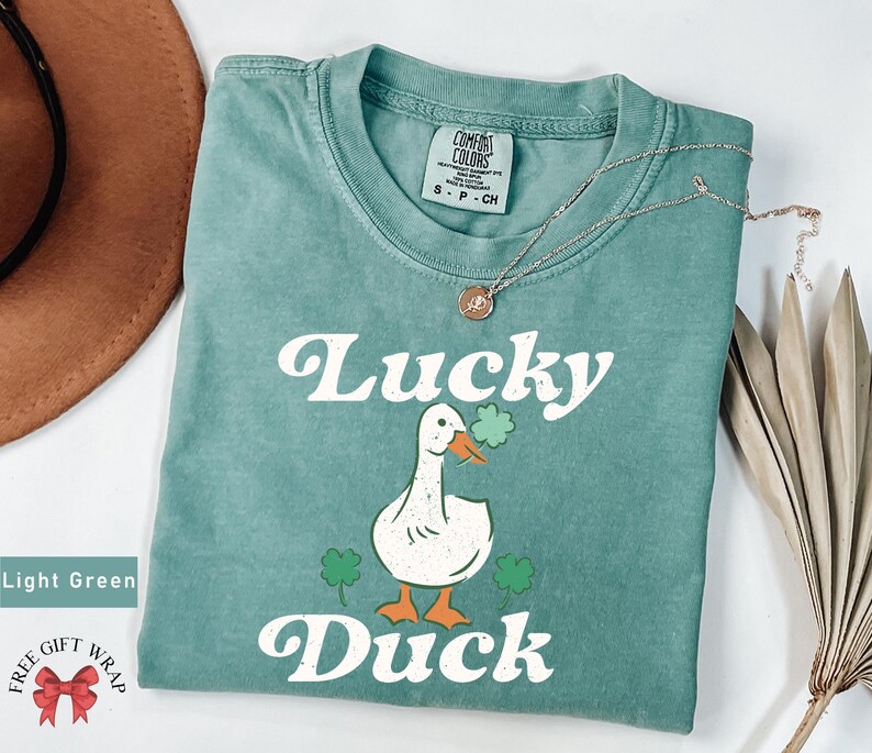 Pu&ograve; includere: Maglietta verde chiaro con un'anatra bianca che tiene un trifoglio e il testo "Lucky Duck" in un font retr&ograve;.