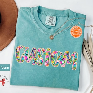 May include: Seafoam green t-shirt with a floral patterned "CUSTOM" design. The shirt is made by Comfort Colors and has a tag that says "S - P - CH".