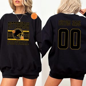 May include: Black sweatshirt with a gold and black Pittsburgh Football design on the front. The design includes a football helmet, the text "PITTSBURGH", "1933", and "FOOTBALL". The back features "CUSTOM NAME" and the number "00".