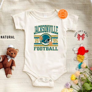 May include: Cream-colored baby onesie with a teal, gold, and white Jacksonville Football design. The design includes a football helmet, the text "JACKSONVILLE", "1995", and "FOOTBALL". The word "NATURAL" is printed on the left side.