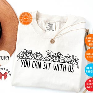 May include: Ivory-colored t-shirt with a black line drawing of people seated at a table, with the text "YOU CAN SIT WITH US". The shirt has a crew neck and short sleeves. A gold necklace is draped on the shirt.