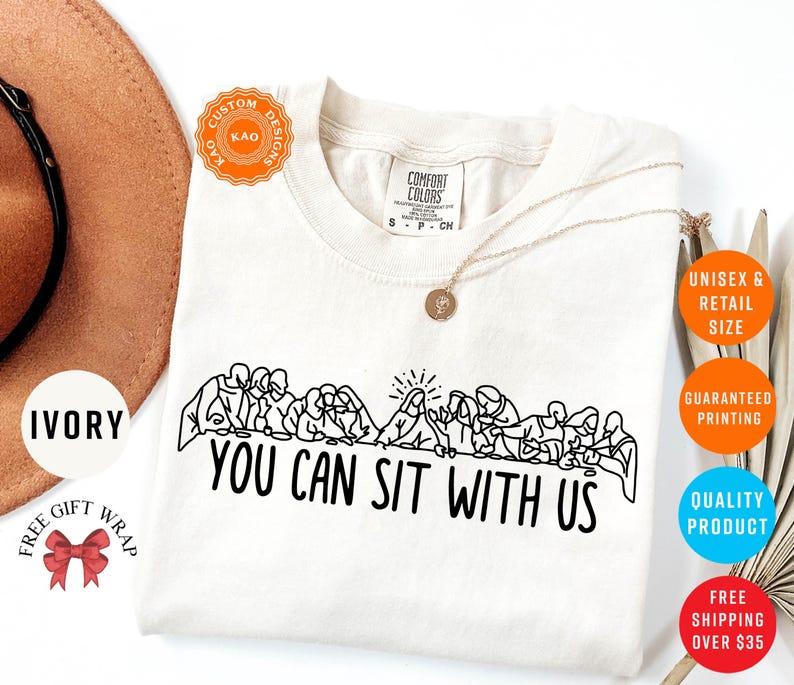 May include: Ivory-colored t-shirt with a black line drawing of people seated at a table, with the text "YOU CAN SIT WITH US". The shirt has a crew neck and short sleeves. A gold necklace is draped on the shirt.
