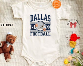 May include: Cream-colored baby onesie with the word "DALLAS" in navy blue block letters above "1960 FOOTBALL". A football helmet graphic is centered between the numbers. The word "NATURAL" is printed on the left side.