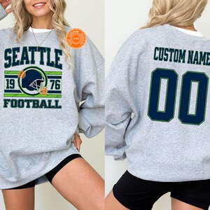 May include: Sport grey sweatshirt with "SEATTLE FOOTBALL 1976" graphic on the front. The design includes a football helmet in navy blue and green. The back of the sweatshirt has "CUSTOM NAME" and the number "00" in navy blue.