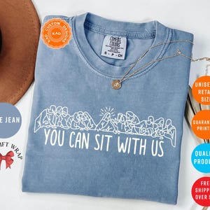 May include: Blue jean colored t-shirt with the phrase "YOU CAN SIT WITH US" in white. The shirt features a line drawing of people sitting together. A gold necklace is draped across the shirt.