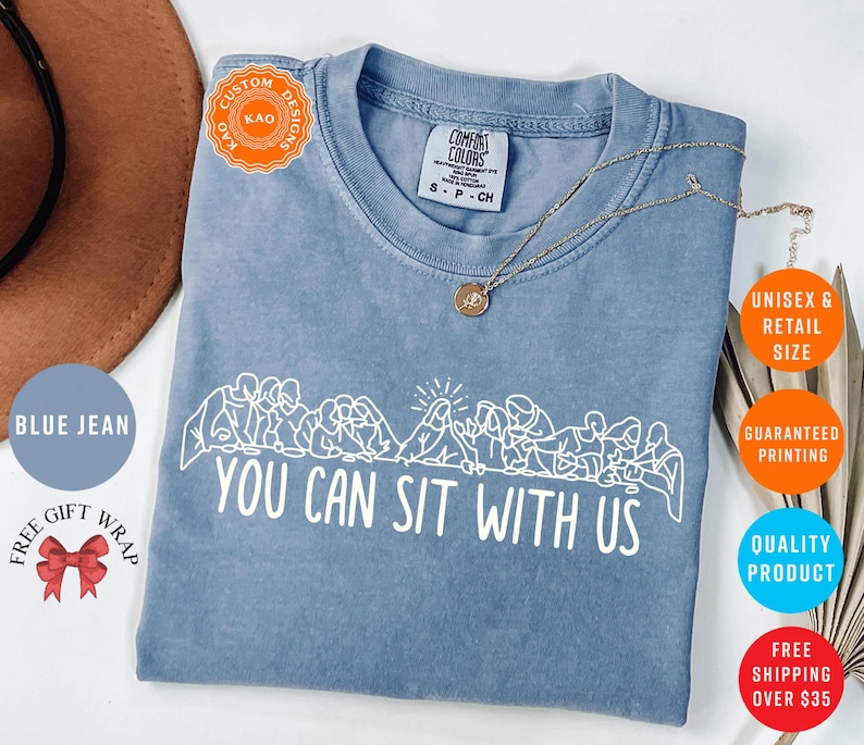 May include: Blue jean colored t-shirt with the phrase "YOU CAN SIT WITH US" in white. The shirt features a line drawing of people sitting together. A gold necklace is draped across the shirt.