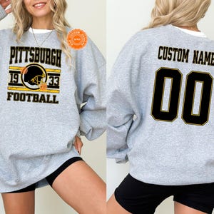 May include: A light gray sweatshirt with the word "PITTSBURGH" in black and gold lettering, along with "1933 FOOTBALL" and a football helmet graphic. The back of the sweatshirt has "CUSTOM NAME" and the number "00" in black and gold.