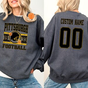 May include: Dark heather gray sweatshirt with "PITTSBURGH FOOTBALL" text and a football helmet graphic in black and yellow. The back features "CUSTOM NAME" and the number "00" in black with yellow trim.