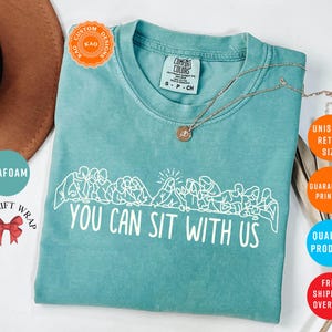 May include: Seafoam green t-shirt with the text "YOU CAN SIT WITH US" in white. The shirt features a graphic of people sitting together. A gold necklace is draped across the shirt. The shirt is a Comfort Colors brand.