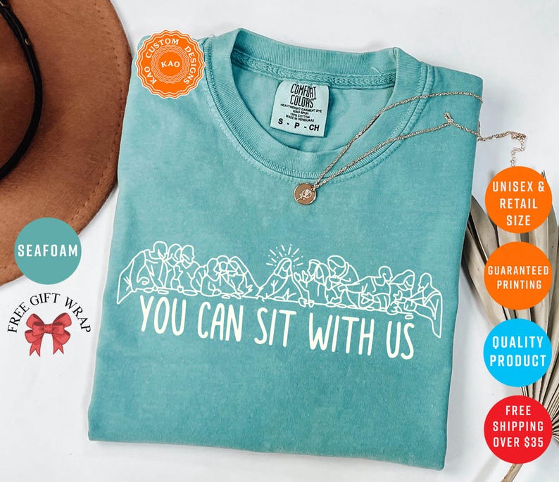 May include: Seafoam green t-shirt with the text "YOU CAN SIT WITH US" in white. The shirt features a graphic of people sitting together. A gold necklace is draped across the shirt. The shirt is a Comfort Colors brand.