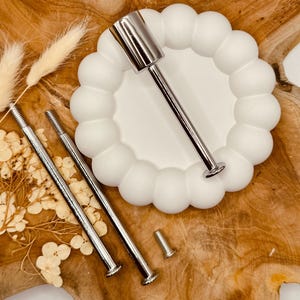 Könnte beinhalten: Ein weißer Keramik-Kuchenständer mit einem runden, gewellten Rand. Der Ständer wird mit drei silbernen Metallstangen und Schrauben zur Montage geliefert.