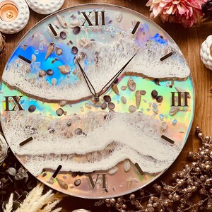 May include: A white and iridescent clock with Roman numerals and a wave design. The clock face is decorated with seashells and small stones.