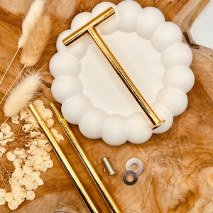 Op de afbeelding: Twee gouden metalen taartstandaards met een T-vormig handvat. De standaards staan op een witte keramische basis met een gegolfde rand. De standaards staan op een houten oppervlak.