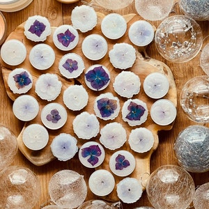 May include: A collection of white votive candles with pressed purple flowers inside. The candles are arranged on a wooden surface with other decorative items.