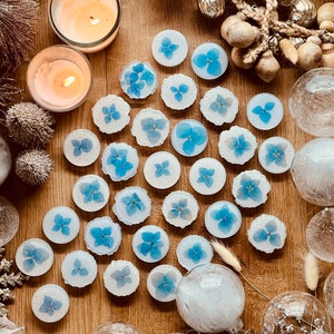 May include: A collection of blue hydrangea flower resin coasters on a wooden surface. The coasters are round and have a white background with a blue flower in the center. There are also several white and clear glass ornaments scattered around the coasters.