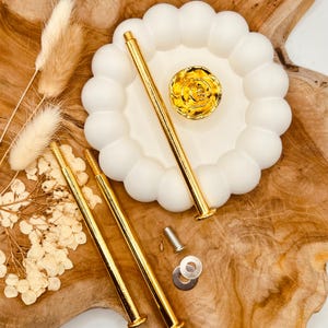 Könnte beinhalten: Drei goldene Metallstangen mit Gewindeenden und einem goldenen, rosenförmigen Dekorationsstück. Die Stangen befinden sich auf einem weißen Keramiksockel mit gewelltem Rand.
