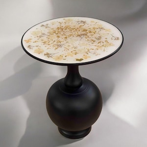 Op de afbeelding: Een ronde zwarte bijzettafel met een wit blad met gedroogde bloemen en gouden bladeren.