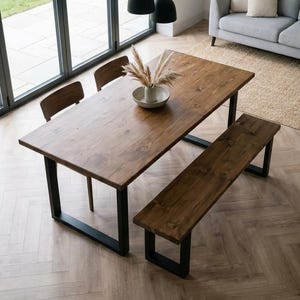 May include: A dining table and bench set with a dark wood finish and black metal frames. The table is surrounded by two wooden chairs. A gray sofa with beige cushions and a woven rug are also in the room. The table is approximately 180 cm long.