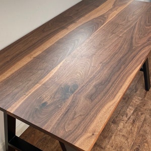 Puede incluir: Una mesa rectangular con un tablero de madera de nogal maciza en color marr&oacute;n oscuro, que muestra vetas y nudos naturales. La mesa se apoya sobre una estructura de metal negro resistente con patas inclinadas para mayor estabilidad.