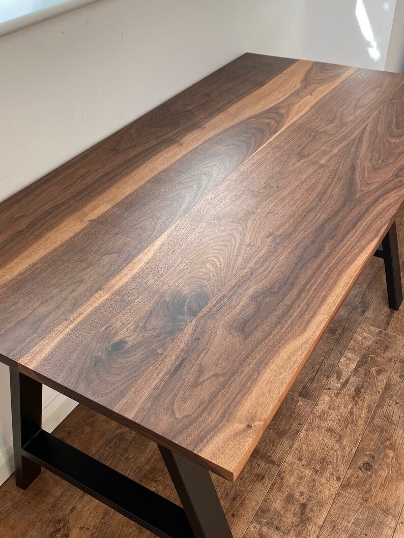Puede incluir: Una mesa rectangular con un tablero de madera de nogal maciza en color marr&oacute;n oscuro, que muestra vetas y nudos naturales. La mesa se apoya sobre una estructura de metal negro resistente con patas inclinadas para mayor estabilidad.